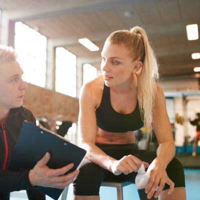 Shot of a personal trainer and young woman discussing fitness plan. Personal trainer showing something on clipboard to student in the gym.
